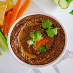 Black bean hummus served with pita chips, celery, and carrot sticks, garnished with olive oil and cilantro.